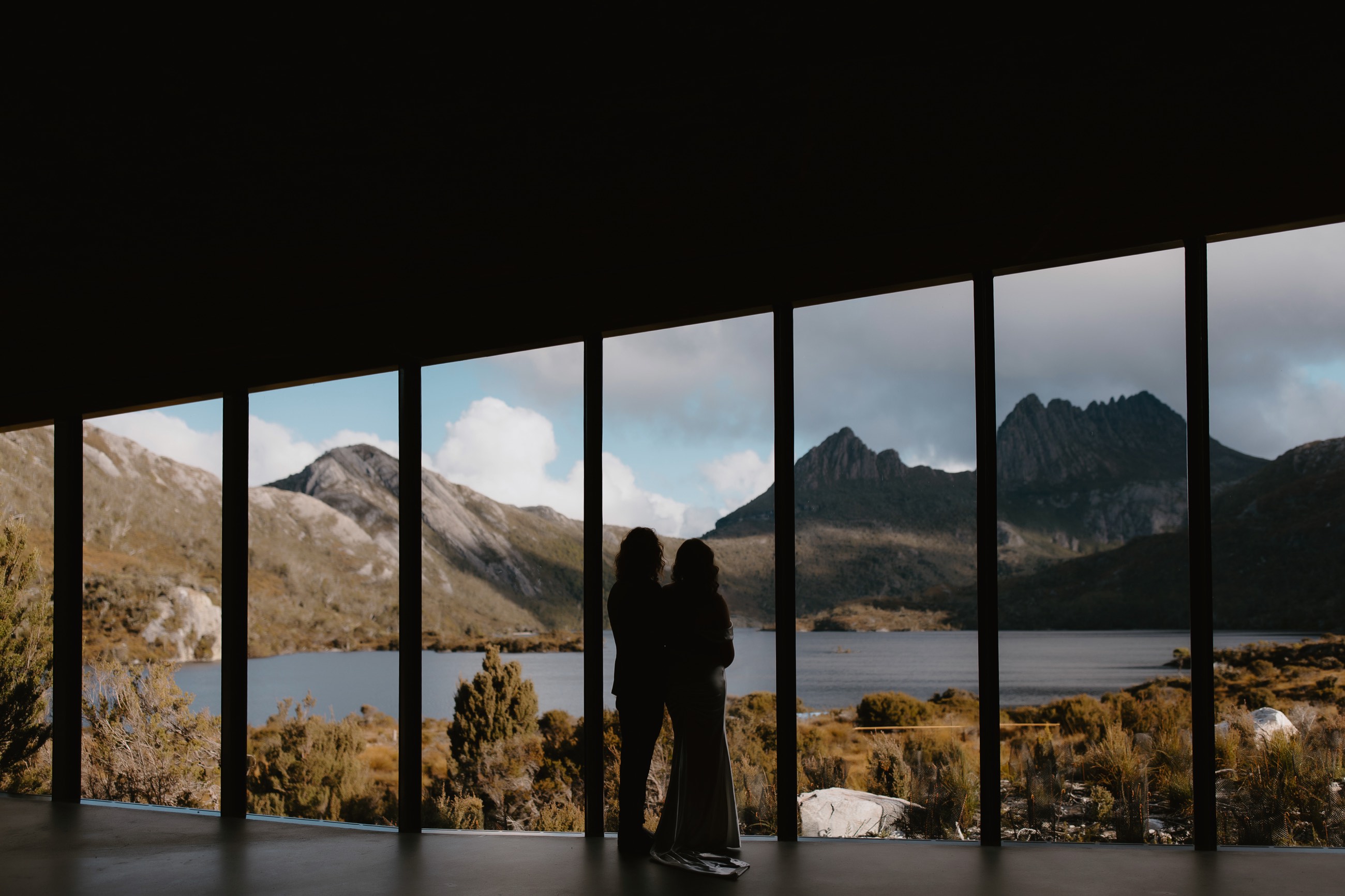 A couple stand together at a wide window looking out to Cradle Mountain and Dove Lake.